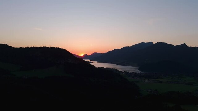 Real-time aerial drone view of serene sunset afterglow over Lake Wolfgangsee and Strobl, Austria, soft pastel sky colors and silhouettes of Salzburg Alps.