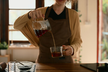 Close up of woman pouring fresh black coffee into a glass cup at home in a warm kitchen
