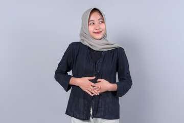 Smiling Asian Muslim woman in casual outfit and hijab posing against a plain background, looking aside with hands on stomach, expressing positive emotion.