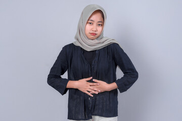 A thoughtful Asian woman wearing a hijab and a dark blouse, gently holding her stomach with both...