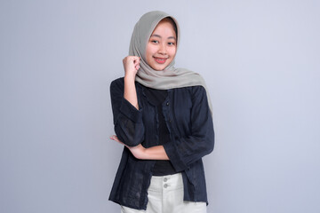 Portrait of a confident young Indonesian Muslim woman wearing a hijab and casual outfit, smiling with a hand on her chin, symbolizing intelligence, strength, and optimism.