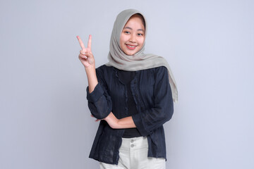 Smiling Indonesian Muslim woman wearing a hijab and black shirt showing a peace sign with her fingers, expressing positivity, harmony, confidence, and friendly communication style.