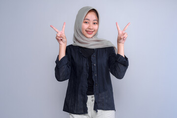 Young Indonesian Muslim woman in hijab smiling joyfully while making double peace signs with both hands, expressing positivity, freedom, cheerful attitude, and youthful energy.