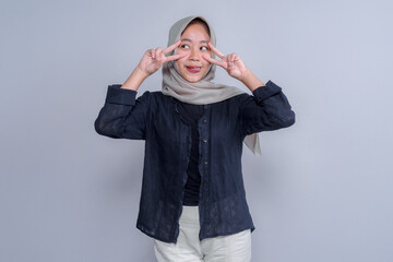 Playful young Indonesian Muslim woman in hijab making double peace signs near her eyes with a funny...