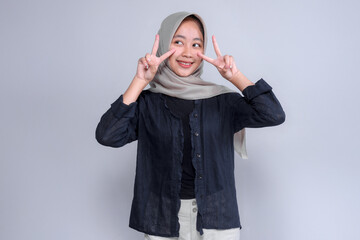 Playful Indonesian Muslim woman in hijab smiling and showing double peace signs near her cheeks, creating a cute and cheerful pose that conveys joy, youthfulness, and positivity.