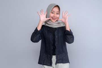 Cheerful young Indonesian Muslim woman wearing a grey hijab and black blouse smiles brightly while playfully raising both hands in front of a plain studio background.