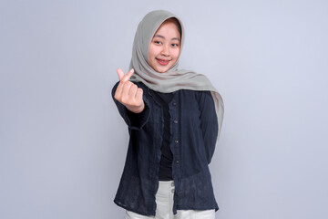Confident young Indonesian woman in a grey hijab and black blouse smiling while making a Korean...