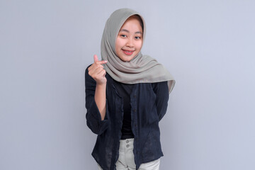 Smiling young Indonesian Muslim woman in a grey hijab and black blouse making a finger heart...