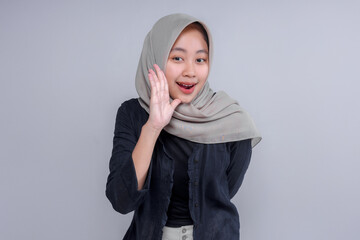 A smiling Asian woman wearing a hijab, posing with one hand raised to her mouth, as if whispering....