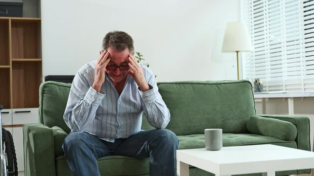 Elderly man sitting on sofa, holding head in hands, showing stress, depression, and loneliness in a home care setting.