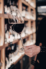 Close-up of a hand holding a glass of red wine in a wine store, with blurred bottles on shelves in the background. Elegant, upscale atmosphere, ideal for lifestyle themes.