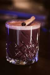 A glass of rich hot chocolate topped with foam and a cinnamon stick, placed on a dark wooden surface under moody lighting.