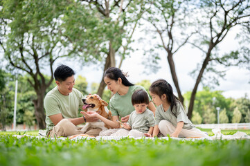 Dad mom feeding corgi puppy while son daughter sitting playing on picnic mat lay over grass in park.