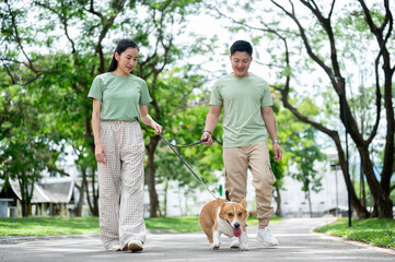 Smiling young asian man and woman holding a leash on corgi puppy while walking on park's pavement.