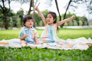 Girl raising hands and boy sibling holding pencil as sitting on picnic mat laying over grass in park
