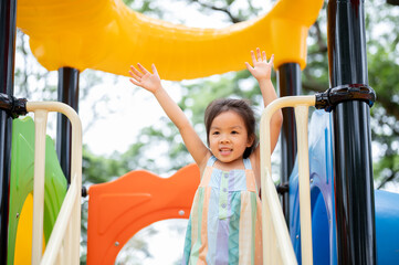 Smiling asian girl standing waving hands on a colorful climbing slide in the park's playground.