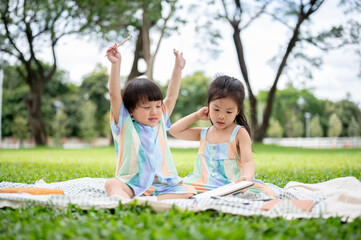 Boy raising hand holding pencil and girl looking at notebook sits on picnic mat over grass in park.
