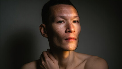 Bare-chested person in soft window light, eyes piercing, skin glowing — quiet vulnerability and strength