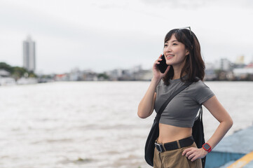 Happy solo Southeast Asian woman teenager blogger on world travel tourism waits for boat using smartphone to express joy to friend by Chao Phraya River in Bangkok Thailand
