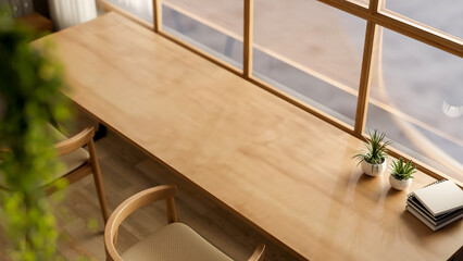 Books and potted plants on long wooden table with armchairs beside glass wall of a cafe or library.