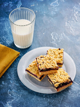 Fresh-baked jam shortbread bars are arranged on a white plate alongside a tall glass of milk. A yellow napkin adds warmth to the inviting kitchen atmosphere