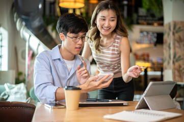 Asian woman bending over table looking at tablet with a man hold calculator sits in cafe or canteen.