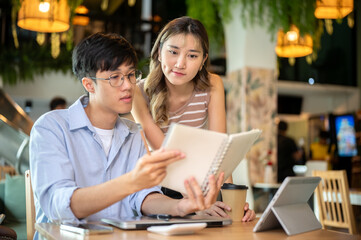 Asian woman standing and reading or looking at notebook in a man's hand at table in cafe or canteen.