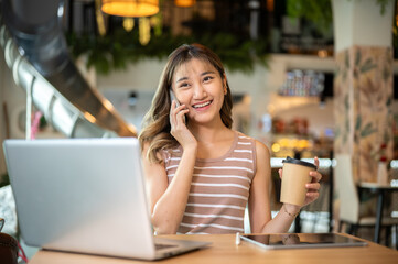 Pretty asian woman holding coffee talking on phone in front of a laptop at table in cafe or canteen.