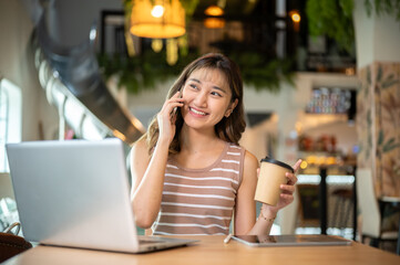 Pretty asian woman holding coffee and talking on phone sits with laptop at table in cafe or canteen.