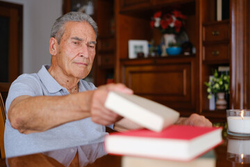 Senior man picking up a book from a table at home, calm indoor scene