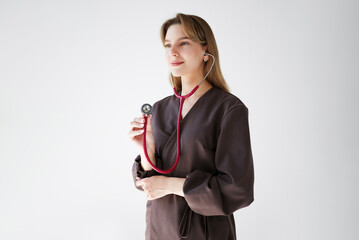 Portrait of a young female doctor in a dark medical uniform listening through a pink stethoscope and smiling slightly, isolated on white background.
