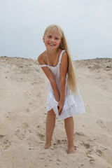 Smiling young blond child girl in white dress standing on sand outdoors