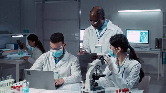 Medium shot of Asian female and Arab male postgraduate students learning to perform blood tests under electronic microscope and record results in data sheet with supervision from Black male professor