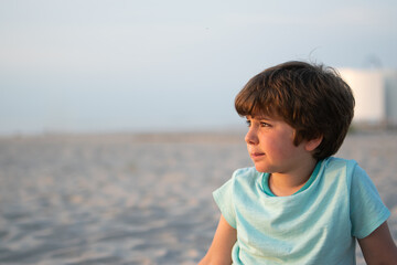 Close-up portrait of a thoughtful boy sitting on a sandy beach during a warm summer evening. Space...
