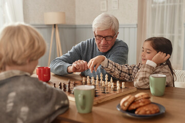 Senior Caucasian man teaching chess to boy and girl at wooden table in home, children concentrating...