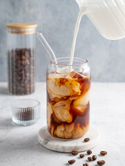 Milk pouring from a milk jug into a glass of iced coffee on a marble board on a light background