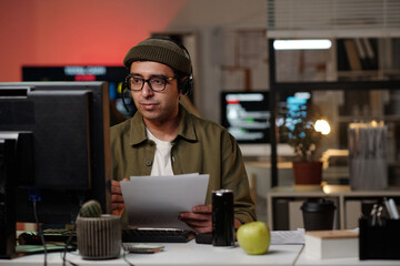 Man wearing headset sitting at desk holding documents working at computer monitors in office...