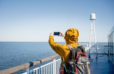 Back view of traveler in jacket taking smartphone photo on ferry deck at sea