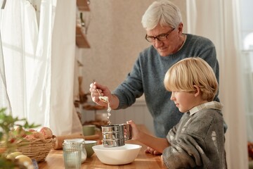 Senior Caucasian man assisting child with baking in rustic kitchen, both standing at wooden table,...
