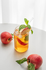 Ice tea with peach, mint in a glass on a white table near window with fresh fruits and sunlight.