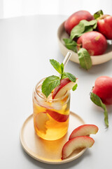 Ice tea with peach, mint in a glass on a white table near window with fresh fruits and sunlight.