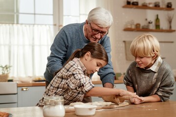 Senior Caucasian man supervising girl and boy making dough together in kitchen, children rolling...