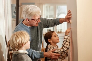 Senior Caucasian man helping girl and boy measuring height on wall in village home, girl holding...