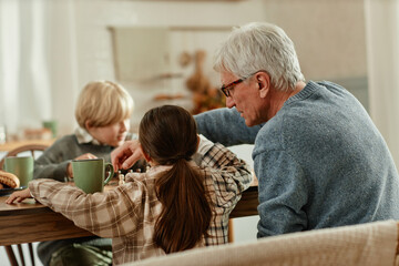 Senior Caucasian man interacting with two children at wooden table, boy and girl sitting beside...