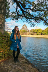 Female tourist in a blue winter jacket stands beside the moat, enjoying the beautiful view of the historic Hiroshima Castle. This peaceful travel scene in Japan is framed by a large, leafy tree