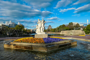 Striking European-style fountain statue creates a fascinating cultural contrast with historic Hiroshima Castle in the background. Water droplets spray in sun, overlooking the moat in Hiroshima, Japan