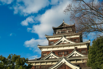 Majestic main keep of Hiroshima Castle showcases its beautiful, traditional architecture against a dramatic blue sky with fluffy white clouds, a famous landmark in Hiroshima, Japan