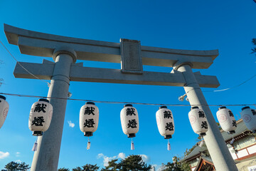 Impressive stone torii gate stands tall against a vibrant blue sky at a Shinto shrine in Hiroshima, Japan. Traditional paper lanterns (chochin) hang in foreground, creating festive, sacred atmosphere