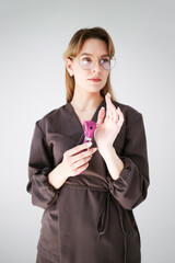 Portrait of a young female doctor in glasses and dark uniform examining her hand with a pink otoscope, isolated on white background.