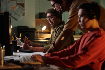 Three young adult men working at desks with headsets in dimly lit office, two sitting and one...